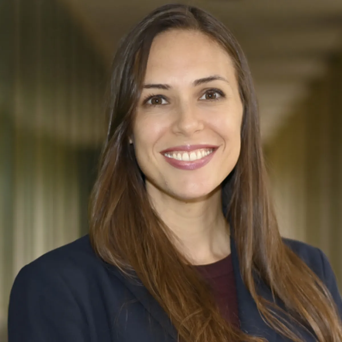 Smiling portrait of a woman with long brown hair wearing dark blue jacket and a maroon top, photographed indoors with a softly blurred background.
